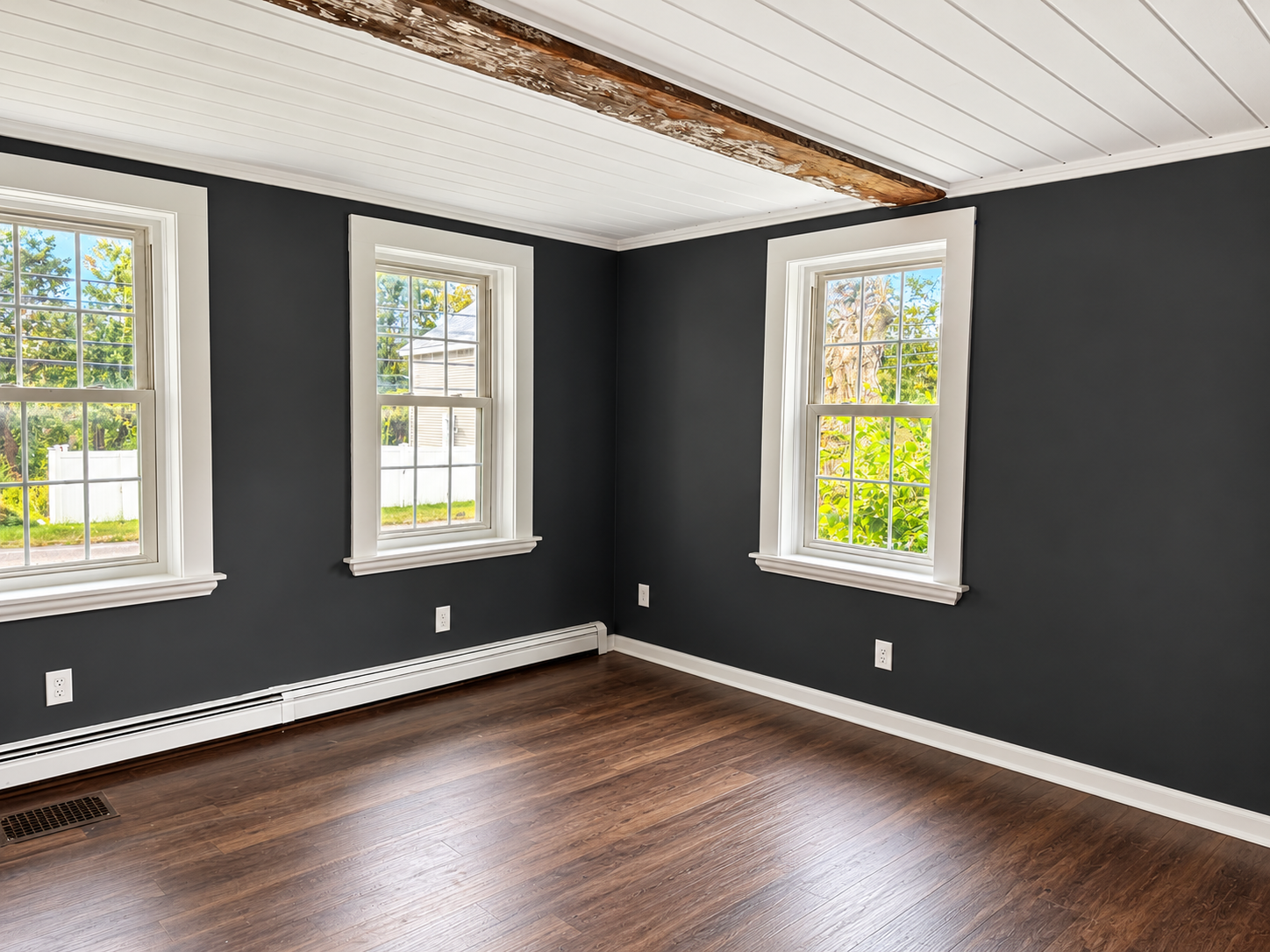 Bright, renovated room featuring charcoal gray walls, white trim, three large windows, dark wood flooring, and a rustic exposed ceiling beam, showcasing the character and natural light of a historic New England home.